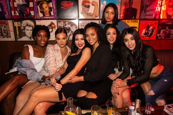 Group of people seated closely together on a couch in a lounge or nightlife setting, with drinks on a table in front and record album covers displayed on the wall behind them