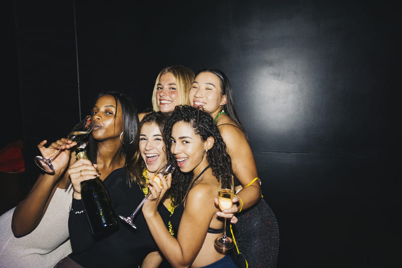 Group of five women smiling and posing with champagne glasses against a black background.