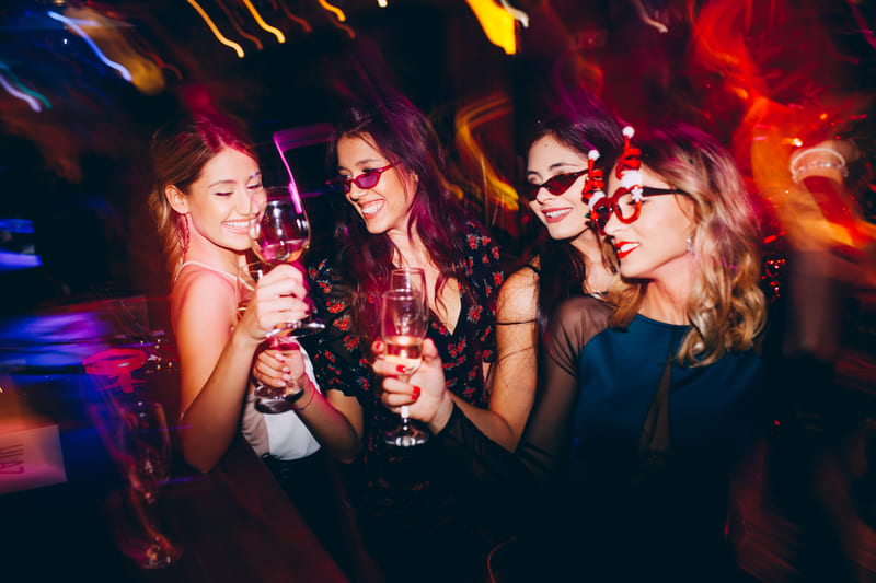 Four women celebrating at a nightclub, wearing fun sunglasses and holding wine glasses under vibrant lights.