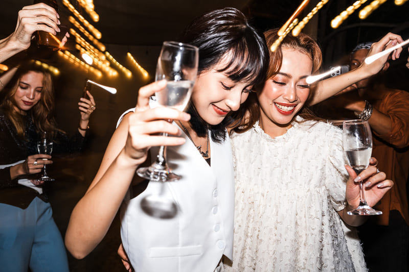 Two women smiling and dancing with drinks in hand at a festive party with string lights.